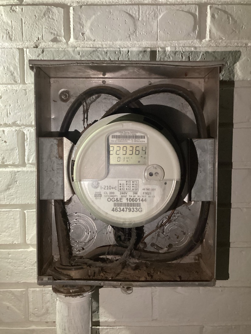 OG&E electric utility meter mounted in exterior box on brick wall.