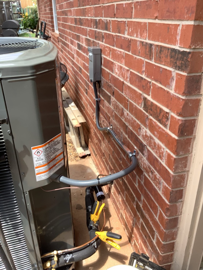 AC condenser unit with refrigerant lines and condensate tank against brick.