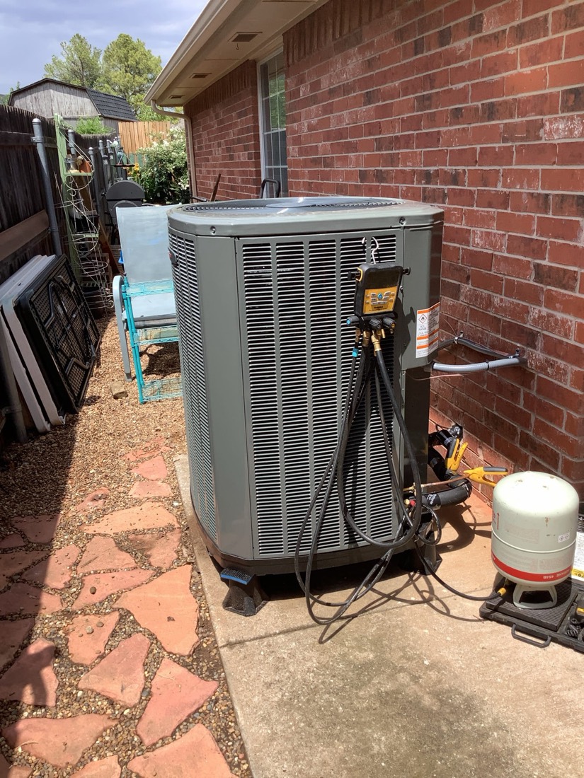 Gray AC condenser unit with hoses and connections on brick foundation.
