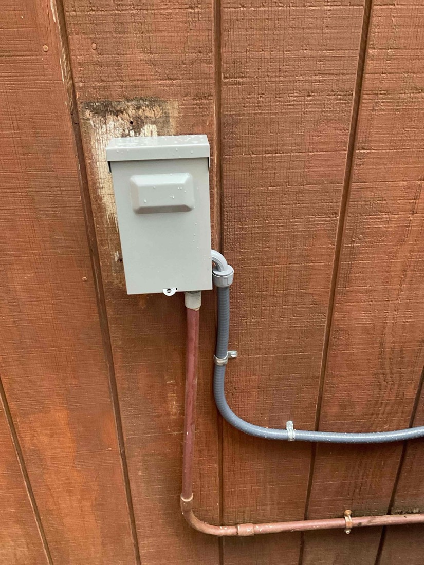 White electrical disconnect switch mounted on wooden fence with copper refrigerant lines.