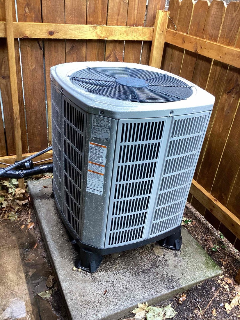 New AC condenser unit installed on concrete pad against wooden fence.