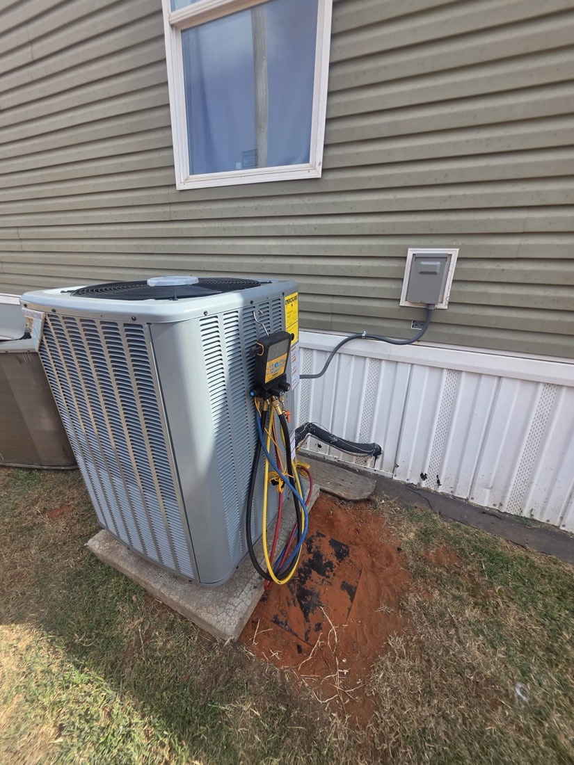 White AC condenser unit mounted on concrete pad with service connections and wall-mounted electrical disconnect.
