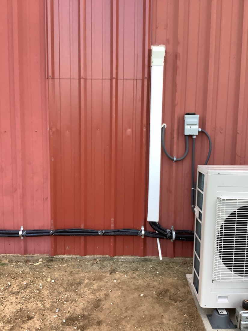 White mini-split air conditioner unit with black conduit and electrical disconnect mounted on red building